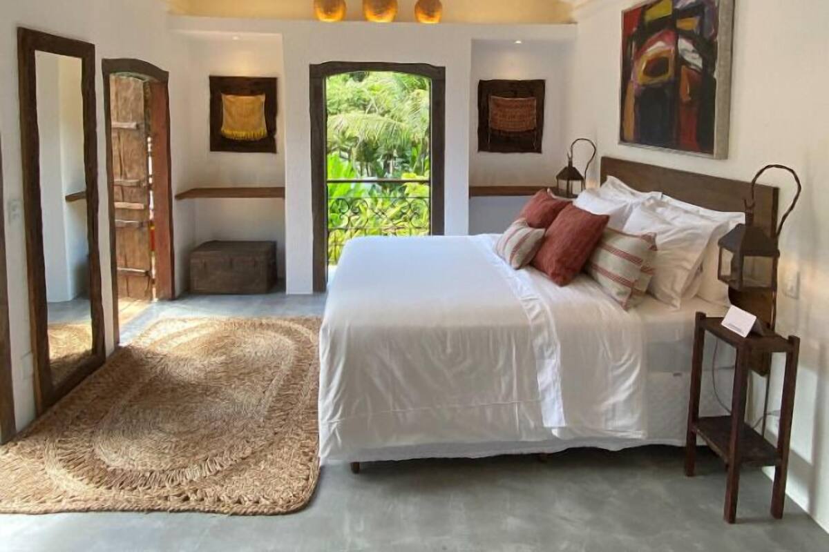 Une chambre avec un grand lit, une tête de lit en bois, un tapis tissé et une vue sur la verdure par une porte.