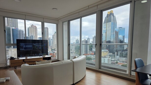 Een moderne woonkamer met een grote televisie, een bank, een eettafel en grote ramen met uitzicht op de stad.