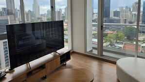 Een moderne woonkamer met een grote flatscreen-televisie, houten vloeren en een stadszicht door grote ramen.