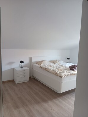 A bedroom with a white bed, a bedside table with a lamp, and a wooden floor.