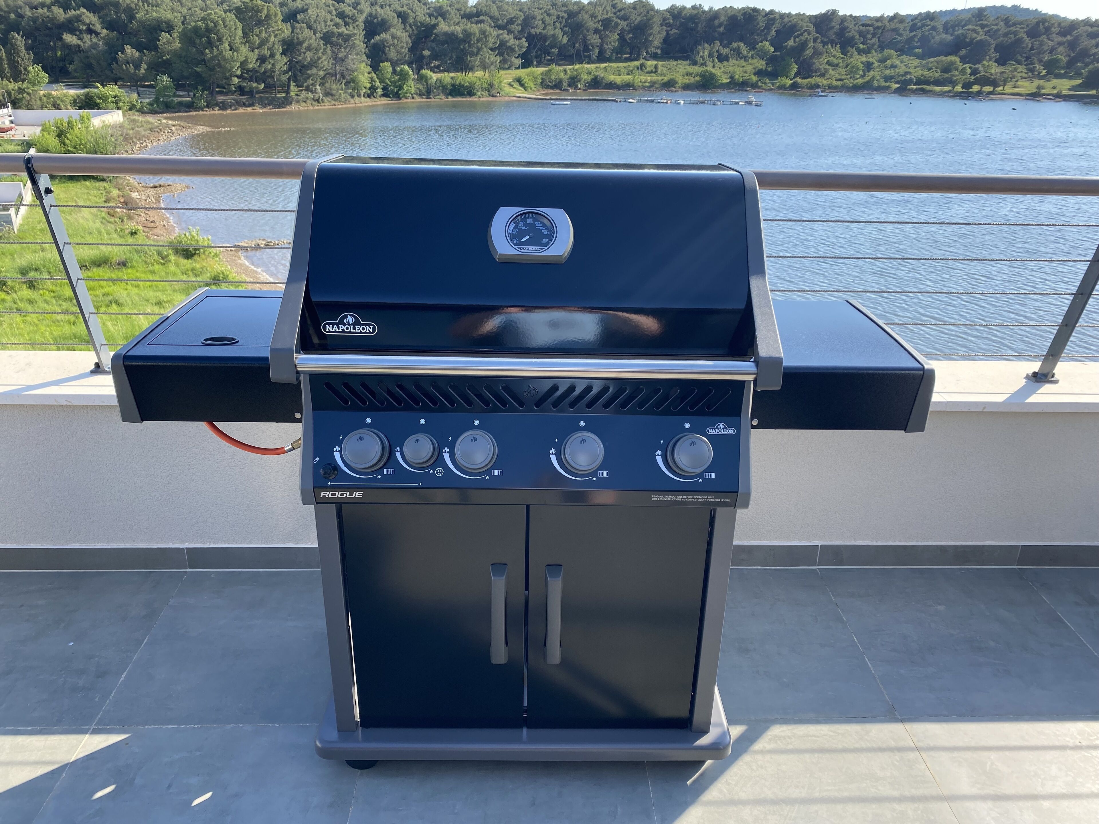 En transportabel gasgrill med fire knapper, placeret på en udendørs terrasse med udsigt til en sø.