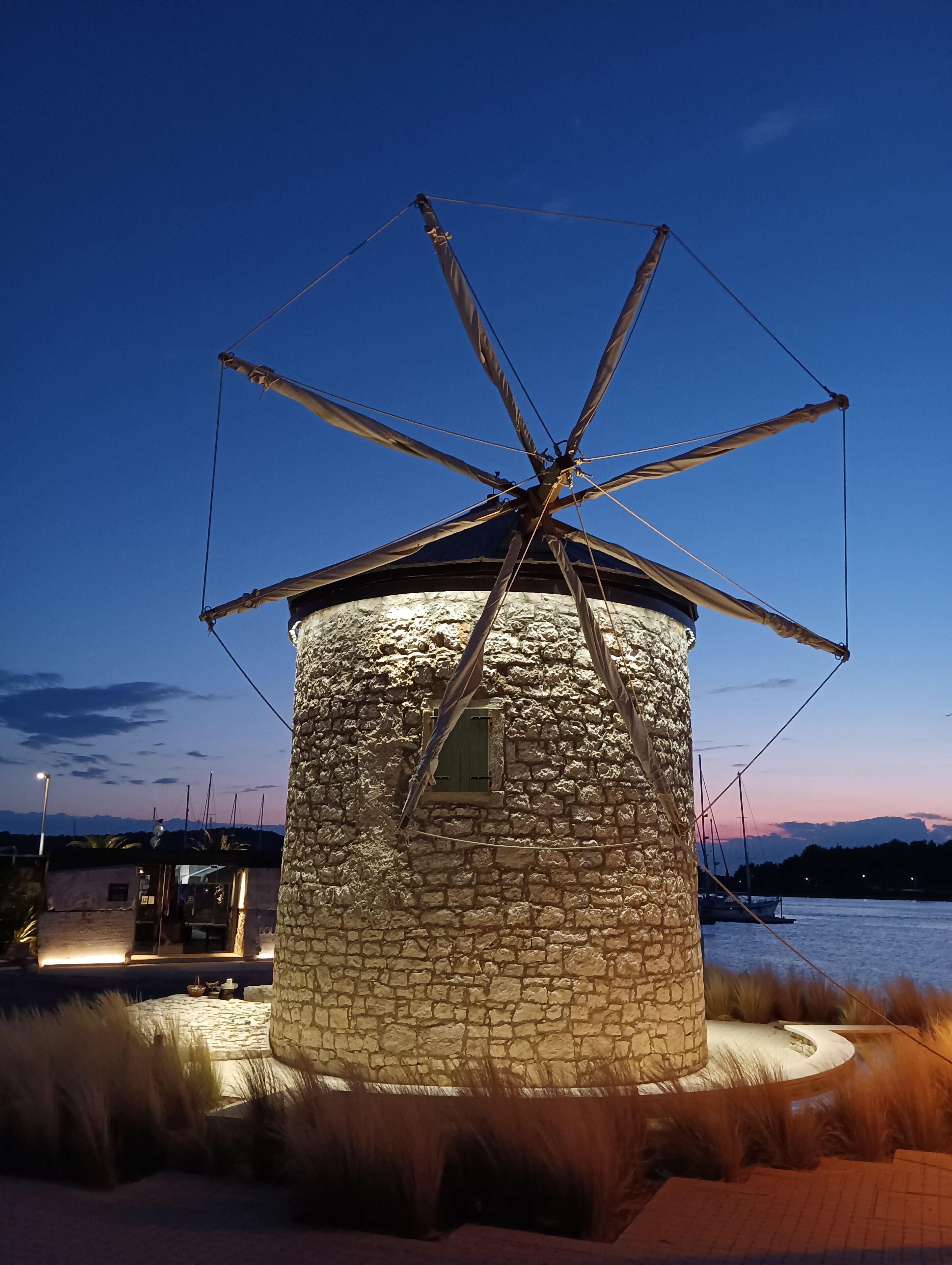 A stone windmill with a metal structure at its top, surrounded by a landscaped area with grass and a body of water in the background.
