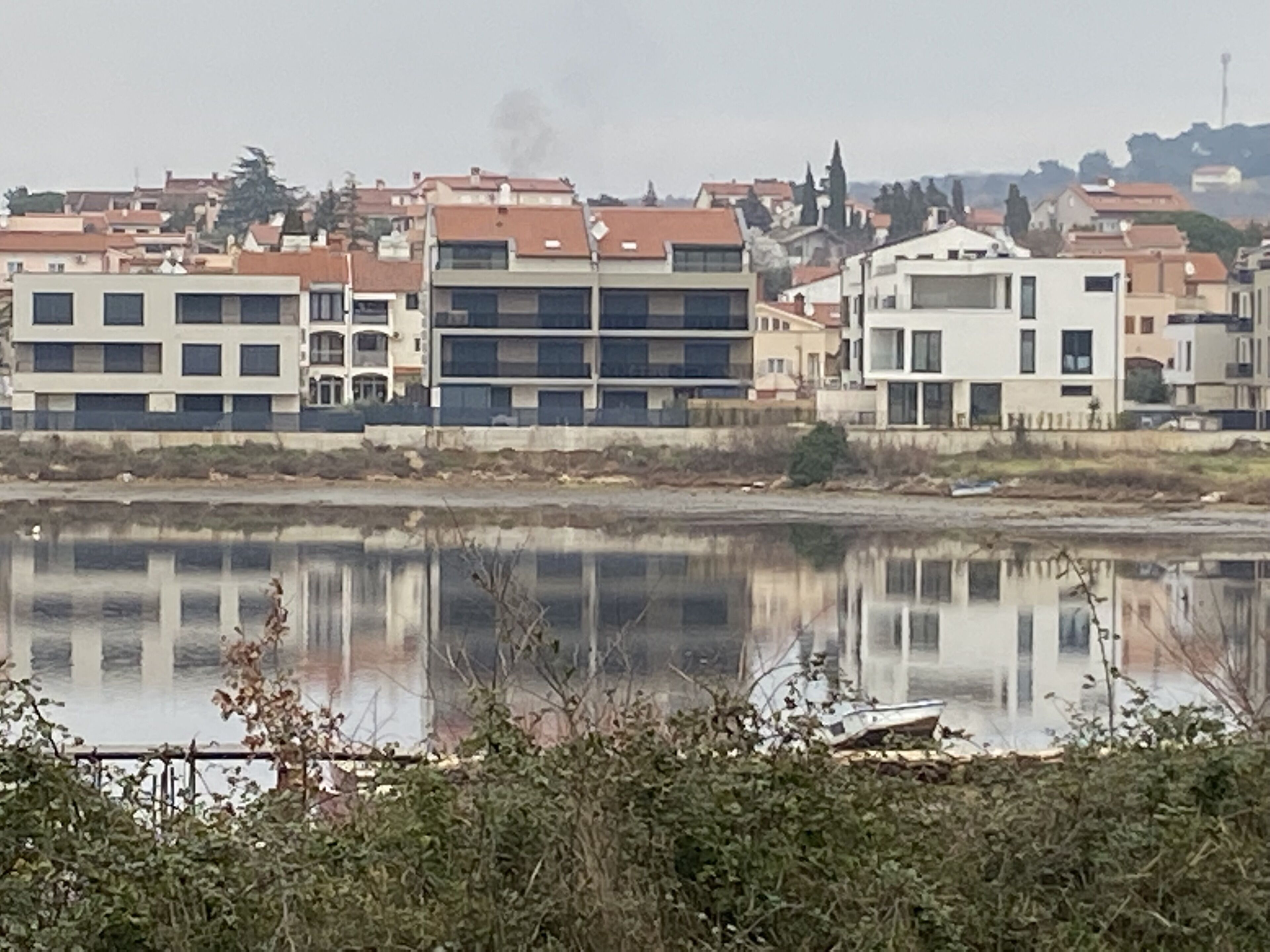 A residential area with modern buildings, a body of water in the foreground, and a hilly landscape in the background.