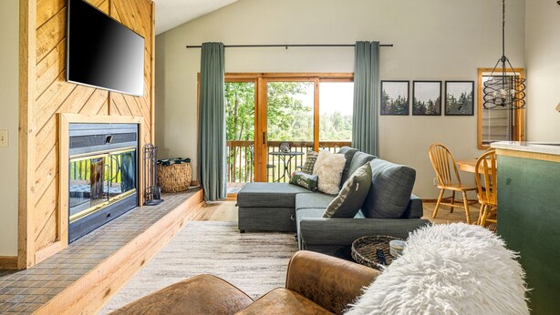 A cozy living room with a fireplace, a sofa, a dining area, and a balcony with curtains. - Northern Michigan paradise!  sleeps 7 KING bed. Private association outdoor pool (Mancelona)