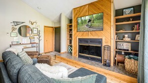 A cozy living room with a fireplace, a mounted TV, and a bookshelf. - Northern Michigan paradise!  sleeps 7 KING bed. Private association outdoor pool (Mancelona)
