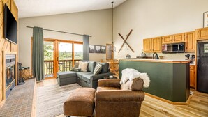A spacious living area with a sofa, ottoman, and a fireplace. A kitchen with wooden cabinets and a large island is visible. - Northern Michigan paradise!  sleeps 7 KING bed. Private association outdoor pool (Mancelona)