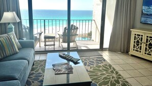 Habitación con sofá, una mesa de centro con control remoto y revistas, vista a la playa y balcón con sillas.
