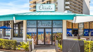 Un restaurante llamado 'Oceans bar & grill' con un toldo turquesa, mesas al aire libre y vista al océano desde la entrada.