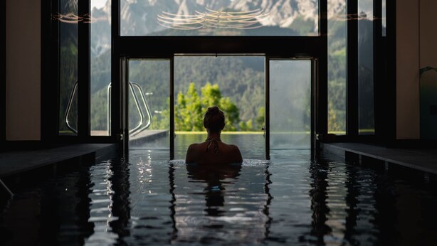 Eine Person im Pool mit Blick auf eine Berglandschaft durch große Fenster.