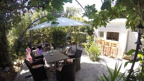 Eine Terrasse im Freien mit Tisch und Stühlen, umgeben von Grünpflanzen und einem weißen Gebäude.
