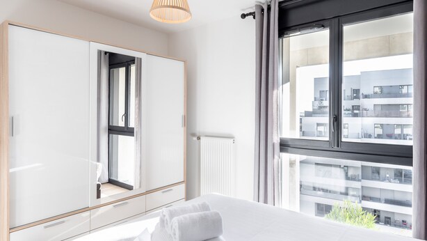 Ein modernes Hotelzimmer mit einem großen Bett, einem Kleiderschrank, einem Fenster mit Blick auf die Stadt und einer Deckenleuchte.