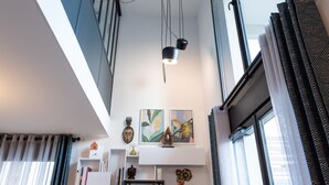 Ein modernes Interieur mit hohen Decken, Pendelleuchten, Kunstwerken und Blick durch ein mit Vorhängen drapiertes Fenster.