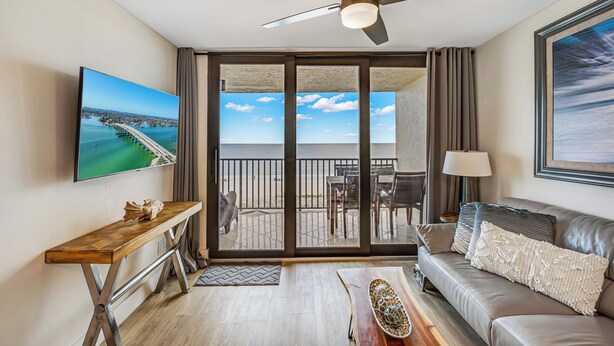 A modern living room with a large TV, a sofa, a coffee table, and a balcony with a view of the beach.
