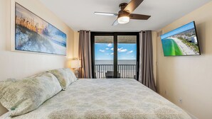 A hotel room with a bed, a ceiling fan, a view of the beach, and two paintings on the walls.