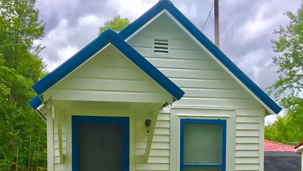 A small, single-story house with a blue door and window, a wooden front porch, and a small garden area.