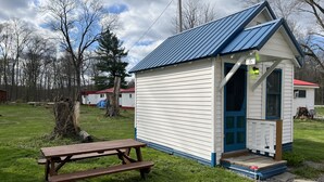 A small, single-story cabin with a blue roof and white siding, a wooden picnic table in front, and a gravel path leading to the entrance.