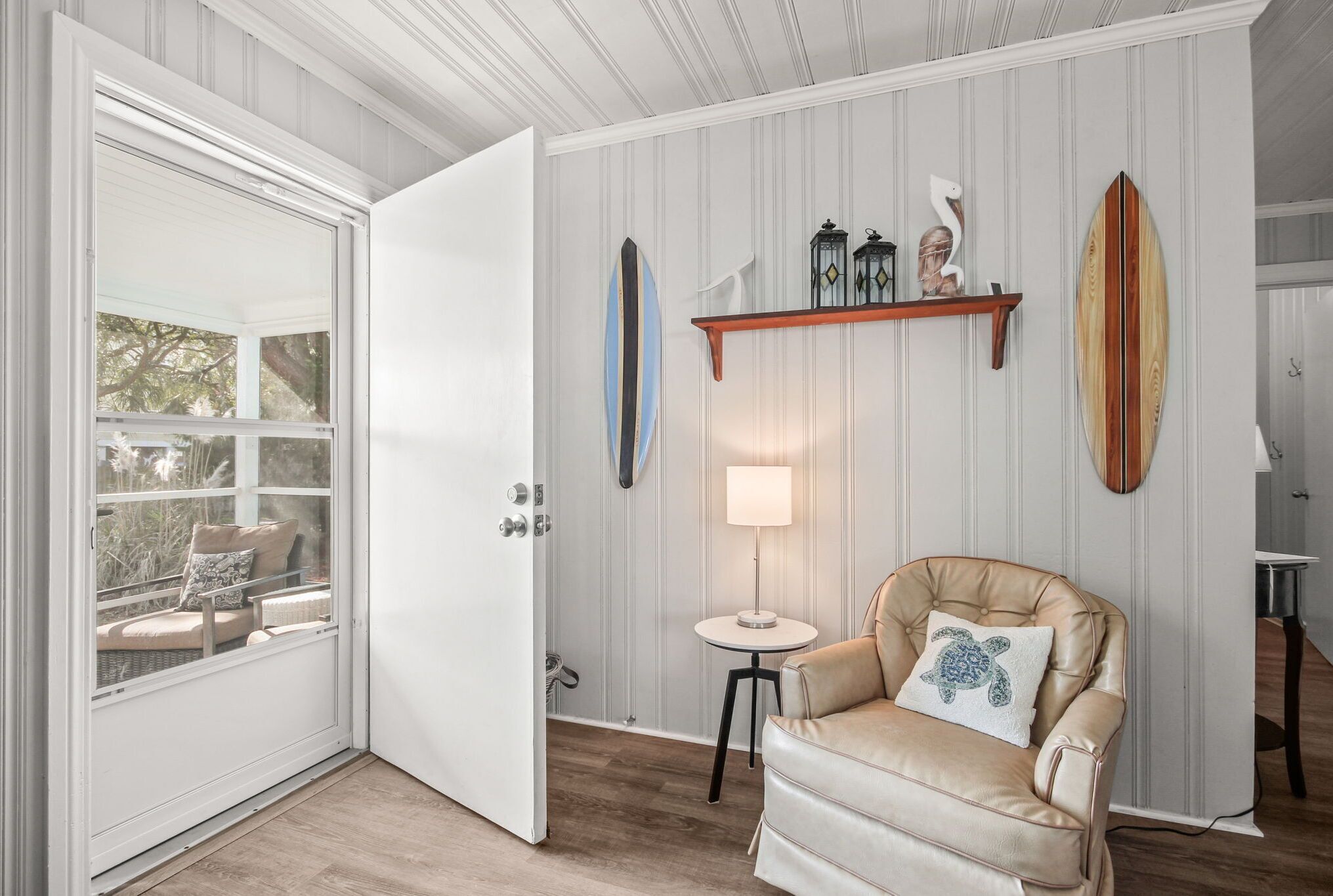 Een kamer met een witte deur, een beige fauteuil met een kussentje in de vorm van een schildpad, een klein rond tafeltje met een lamp, en surfplanken op een plank.