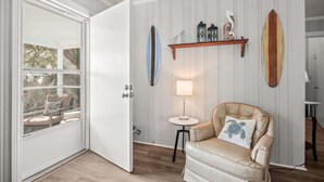 Una habitació amb una porta blanca, un otomà beix amb un coixí amb forma de tortuga, una taula petita i rodona amb una làmpada, i uns surf abocats a l'estant.
