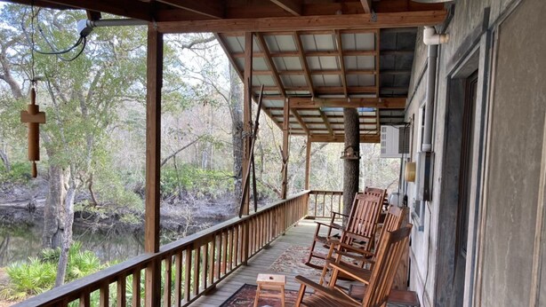 A covered porch with wooden chairs and a small table, overlooking a natural landscape with trees and a body of water. - Skip’s Rustic River Cabin near Okefenokee Swamp (Folkston)
