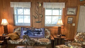 A cozy room with wooden walls, two windows with white curtains, a sofa with a floral print, a wooden coffee table, and two lamps. - Skip’s Rustic River Cabin near Okefenokee Swamp (Folkston)