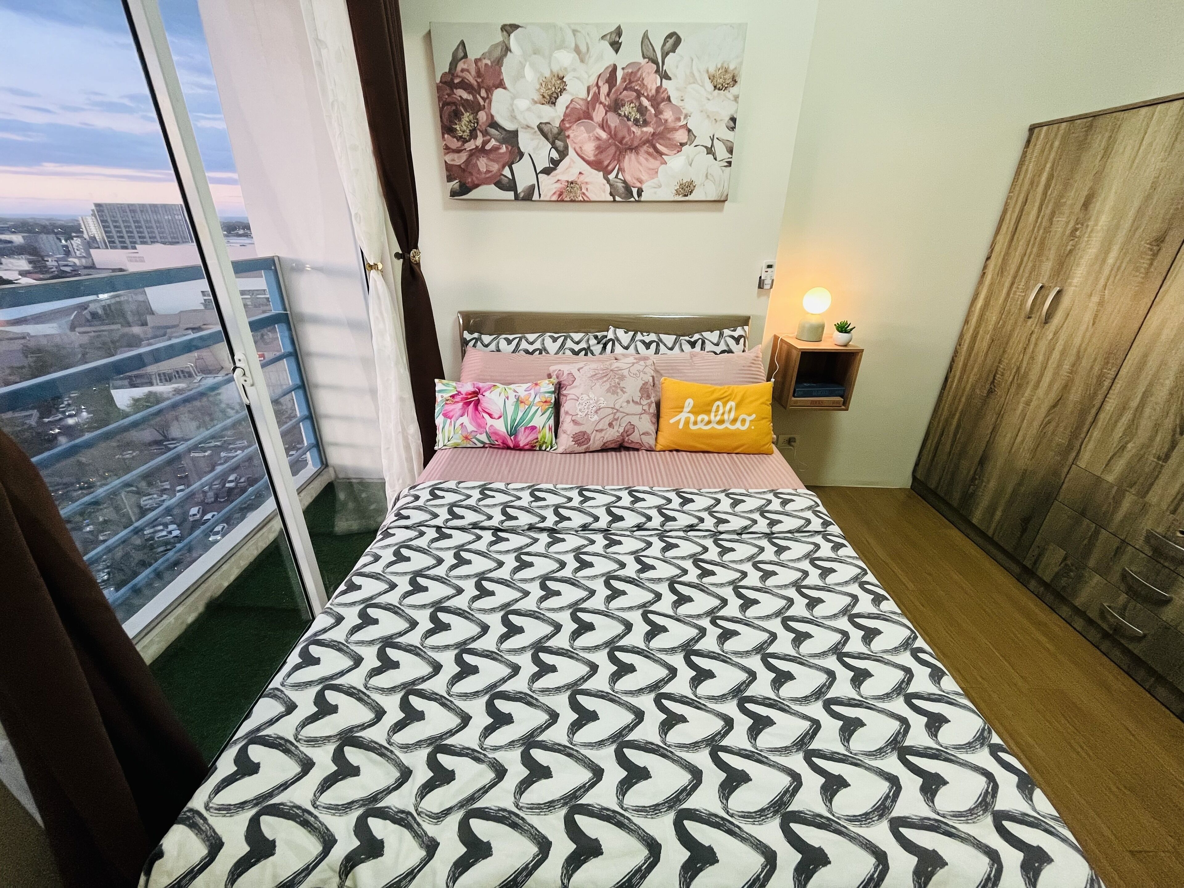 Ein Schlafzimmer mit gemustertem Bettbezug, einem Blumenbild über dem Bett, einem Nachttisch mit Lampe und Blick auf die Stadt vom Balkon.