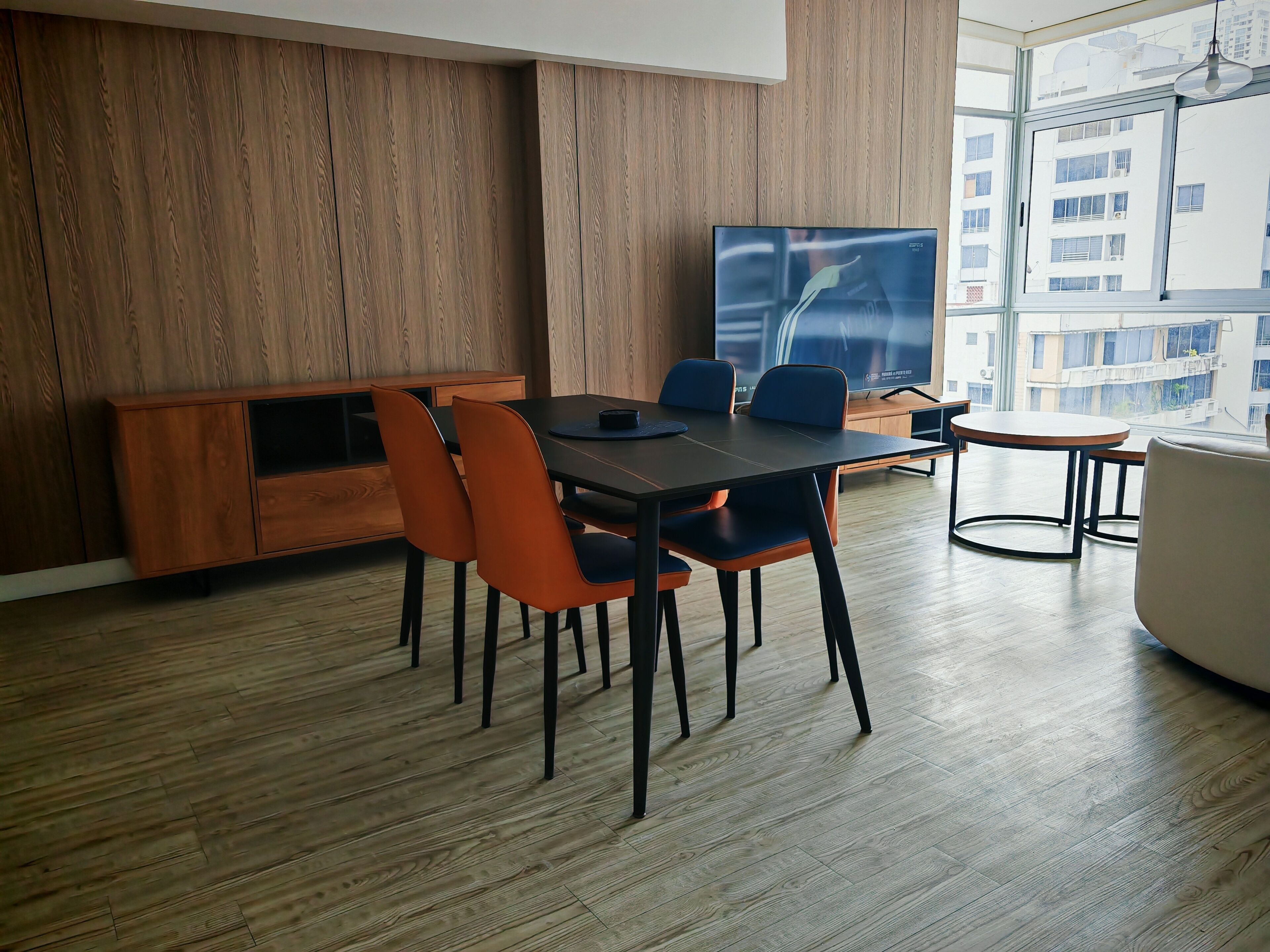 A modern dining area with a wooden wall, a large flat-screen TV, and a view of buildings through large windows.