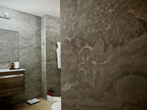 Un baño con pared de mármol, tocador de madera, espejo, lavamanos blanco y toallero.