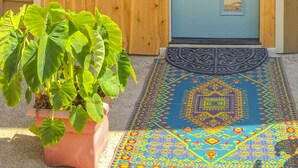 Une maison avec une clôture en bois, une porte bleue ornée de petites fenêtres et un tapis coloré devant l’entrée.