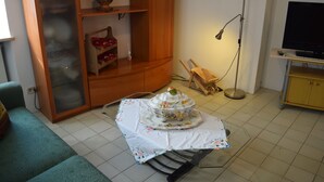 Un salon avec une table à manger recouverte d’une nappe à motifs floraux, un buffet en bois avec des portes en verre, un téléviseur et un lampadaire.
