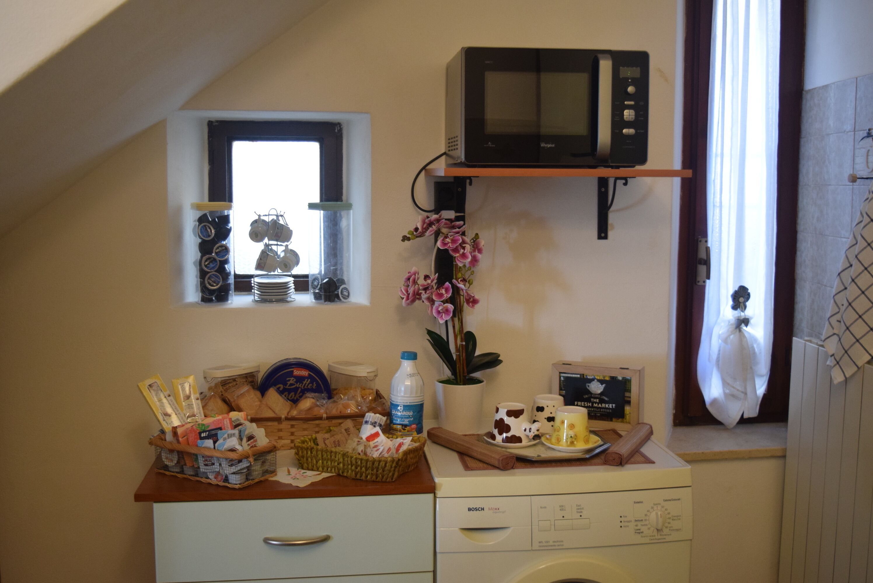 Ett kök med en mikrovågsugn, ett fönster med upphängda köksredskap, en diskho och en bänkskiva med matvaror samt en tvättmaskin.