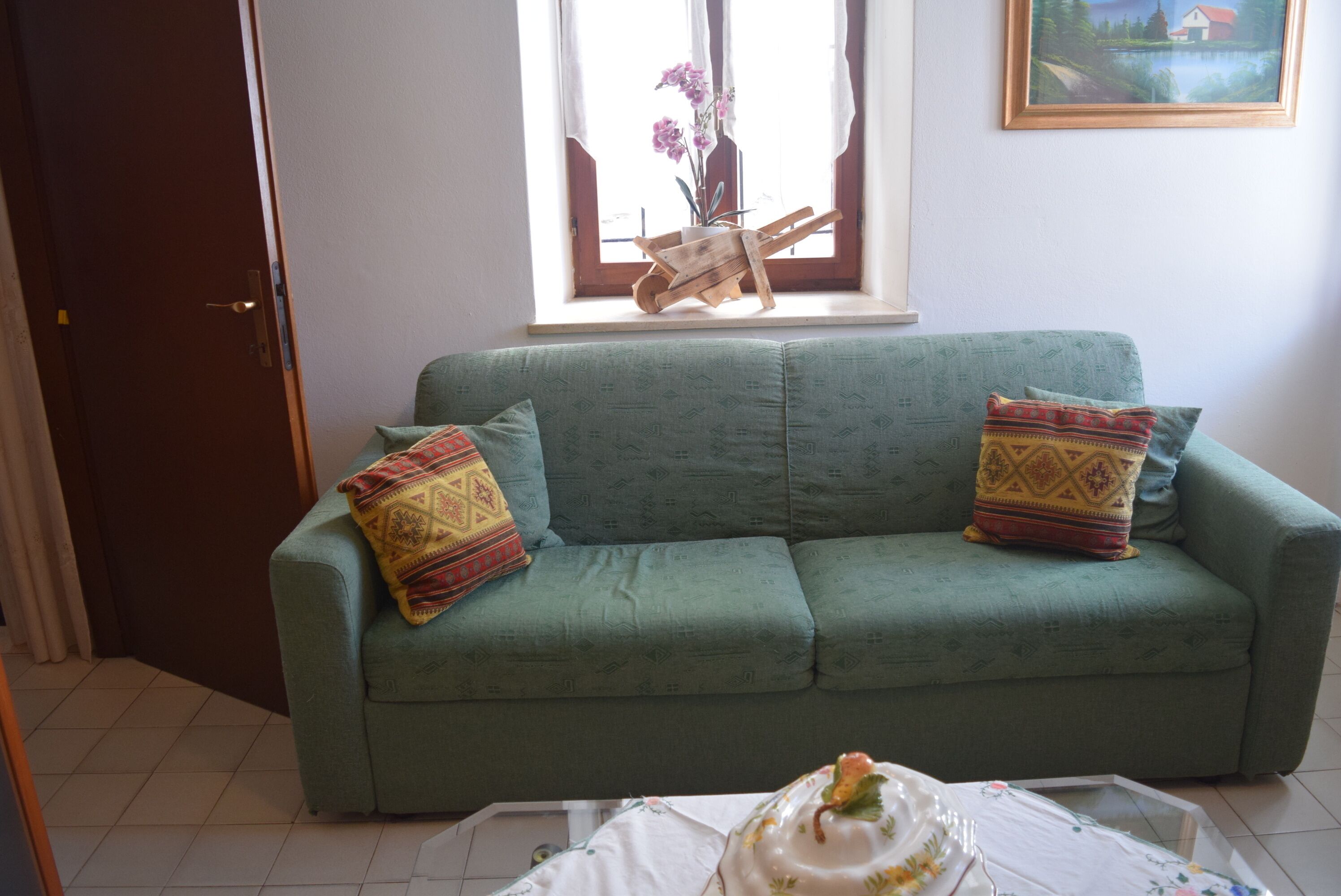 Ein grünes Sofa mit gemusterten Kissen, ein Holzstuhl und eine Vase mit Blumen auf einem Fensterbrett.