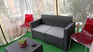 Un canapé en osier avec des coussins gris, deux chaises rouges, une petite table avec une plante et une bougie, et un sol recouvert d’un gazon synthétique vert.