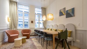Een eethoek met een houten tafel, zwarte stoelen en een groene kruk. Er staan twee roze fauteuils en een vloerkleed met een patroon.