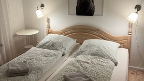 Ett sovrum med en sänggavel i trä, två sängar med vita, strukturerade sängkläder, en grå handduk på varje säng, ett väggmonterat konstverk föreställande ett rådjurshuvud och två väggmonterade lampor.