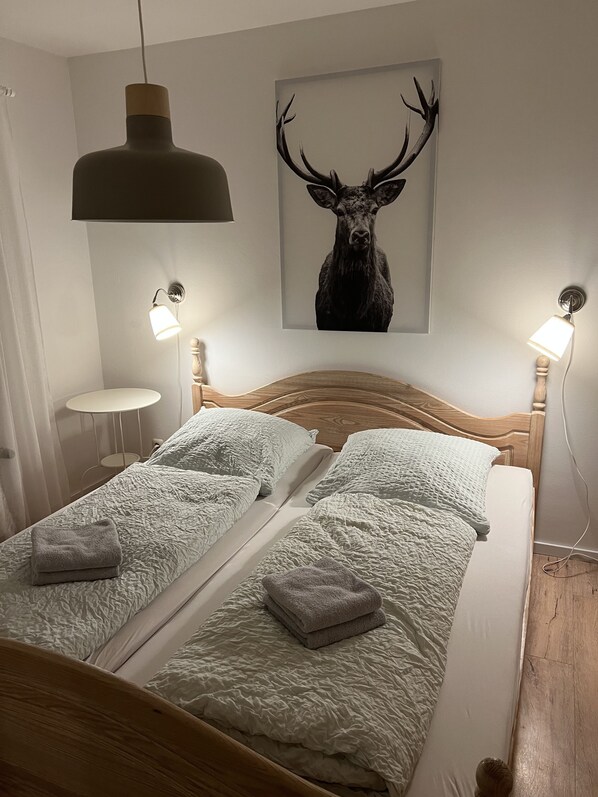 Une chambre avec une tête de lit en bois, deux lits avec une literie blanche à texture, une serviette grise sur chaque lit, une décoration murale représentant une tête de cerf, et deux lampes fixées au mur.