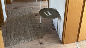En hall med en dörr av trä, en vit dörr, ett litet bord med ett tidningsnummer och en vit stege som står lutad mot väggen.