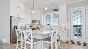 A modern kitchen with white cabinetry, a stainless steel refrigerator, a marble island with bar stools, and a view of the outdoors through large windows. - A FEW STEPS TO OCEAN in EMERALD ISLE, NC! Come visit walk to all entertainment! (Emerald Isle)