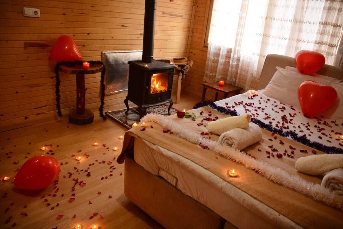A cozy room with a wood-burning stove, a bed with rose petals and heart-shaped balloons, and a wooden interior.