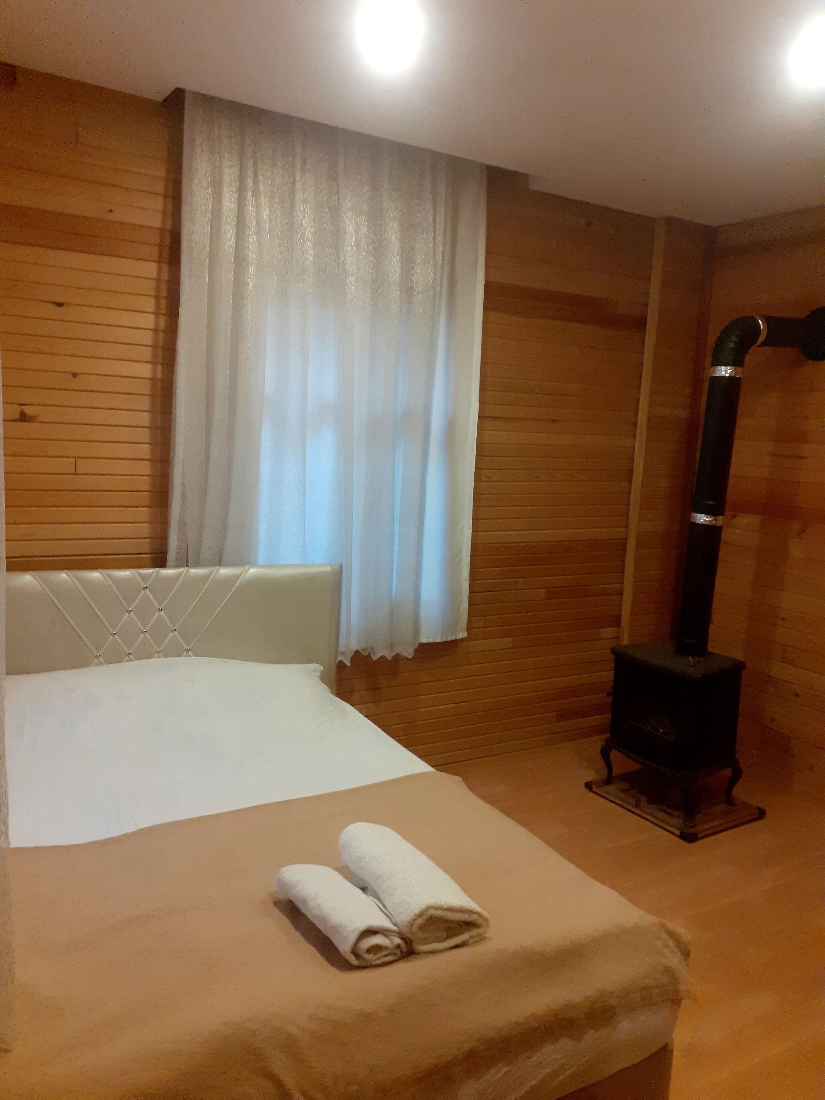 A room with a bed, a wooden headboard, a white curtain, a wooden wall, and a black heater.