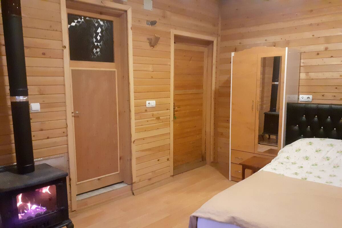 A wooden cabin interior with a wood-burning stove, a bed, a mirror, and a wardrobe.