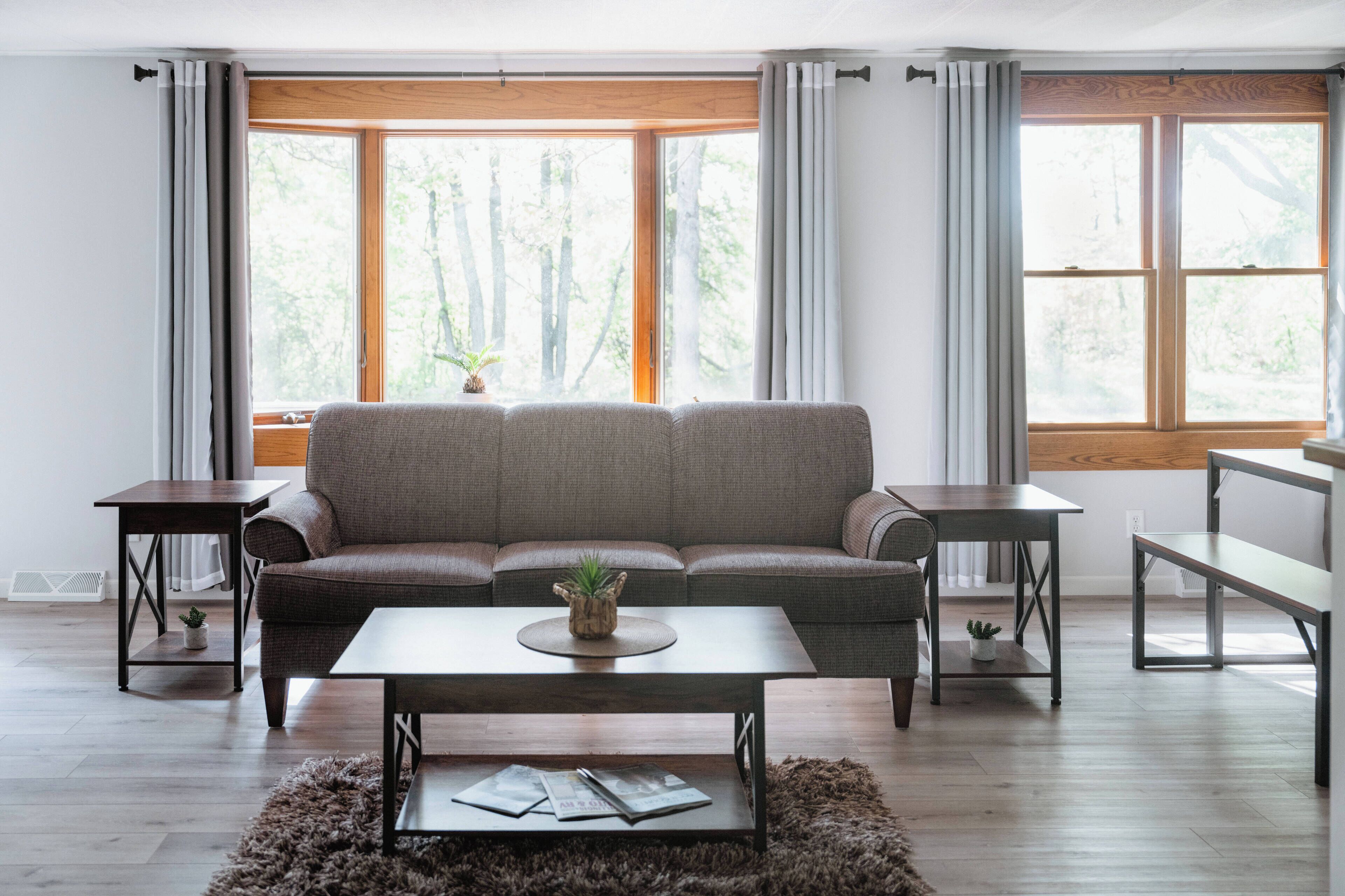 Una sala de estar con un sofá, una mesa de centro y dos mesitas auxiliares. Hay ventanas grandes con cortinas, una pequeña planta sobre la mesa de centro y una alfombra en el suelo.