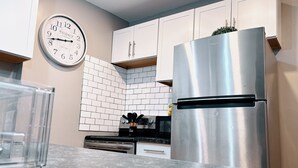 Een keuken met witte kasten, een roestvrijstalen koelkast, een klok aan de muur en een betegelde spatwand.