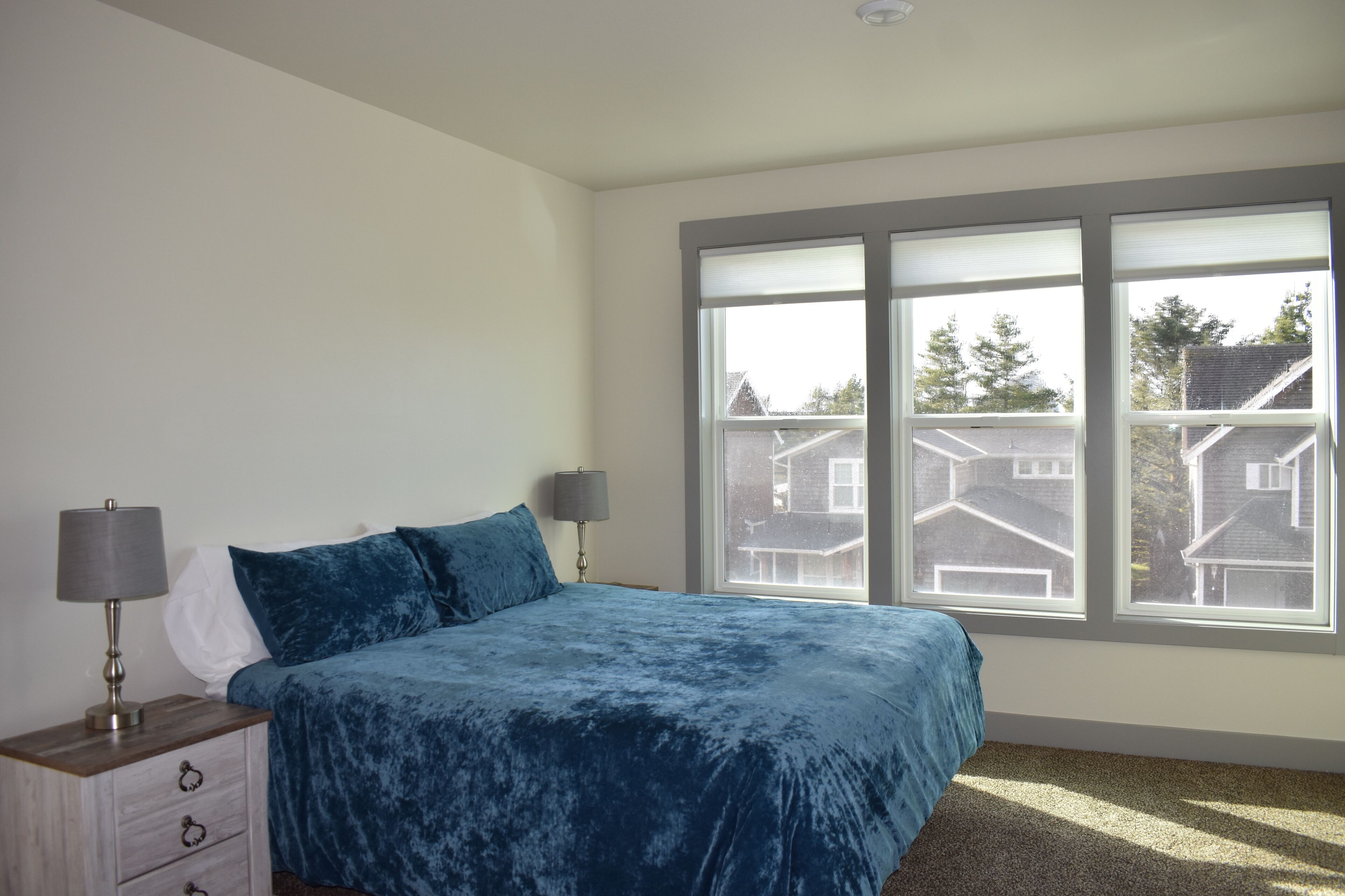 Um quarto com uma janela grande, uma cama com colcha azul, duas luminárias de cabeceira e uma mesa de cabeceira com gavetas.