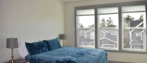 Um quarto com uma janela grande, uma cama com colcha azul, duas luminárias de cabeceira e uma mesa de cabeceira com gavetas.
