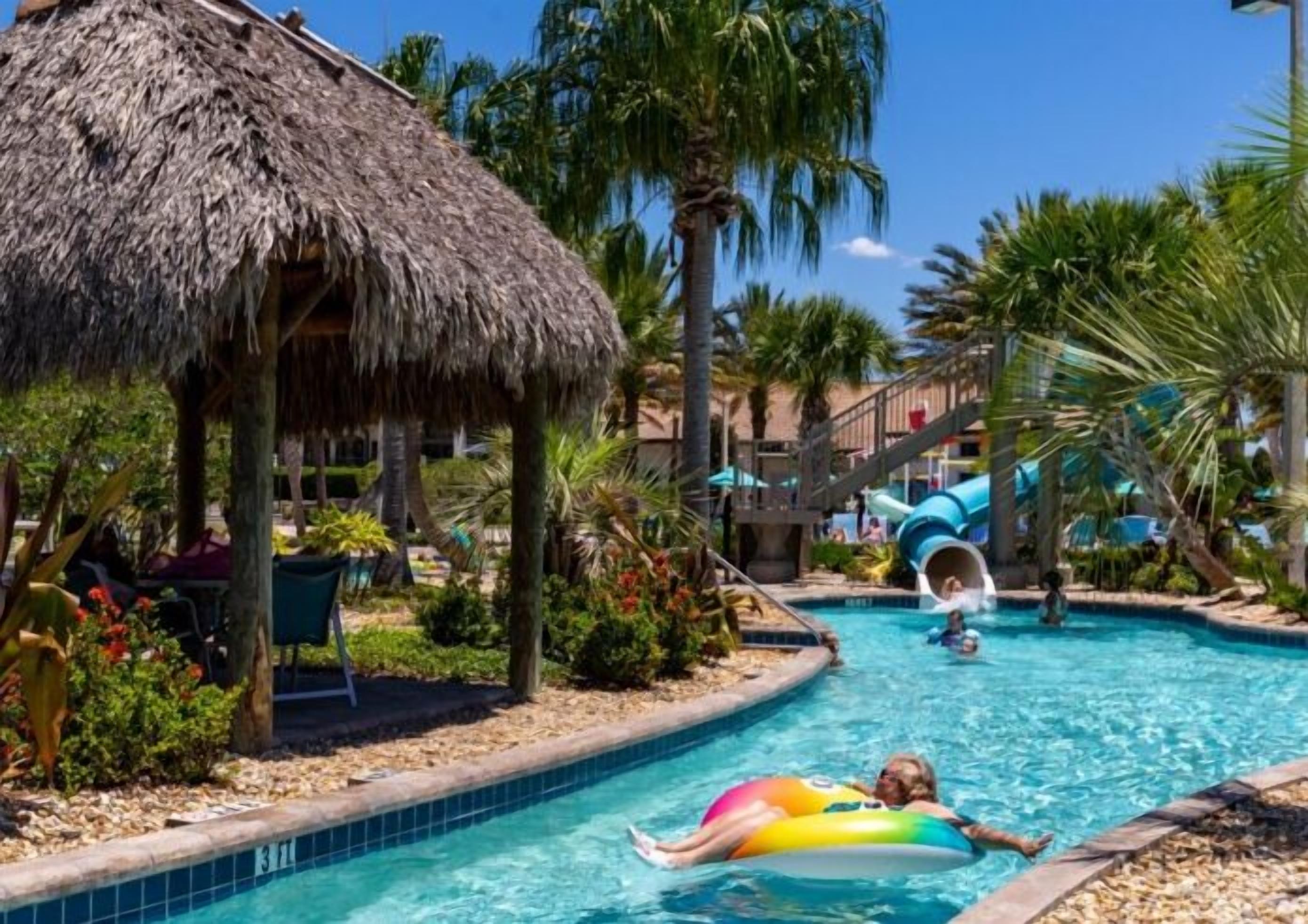 A pool area with a thatched-roof structure, a water slide, and a person floating on an inflatable ring.