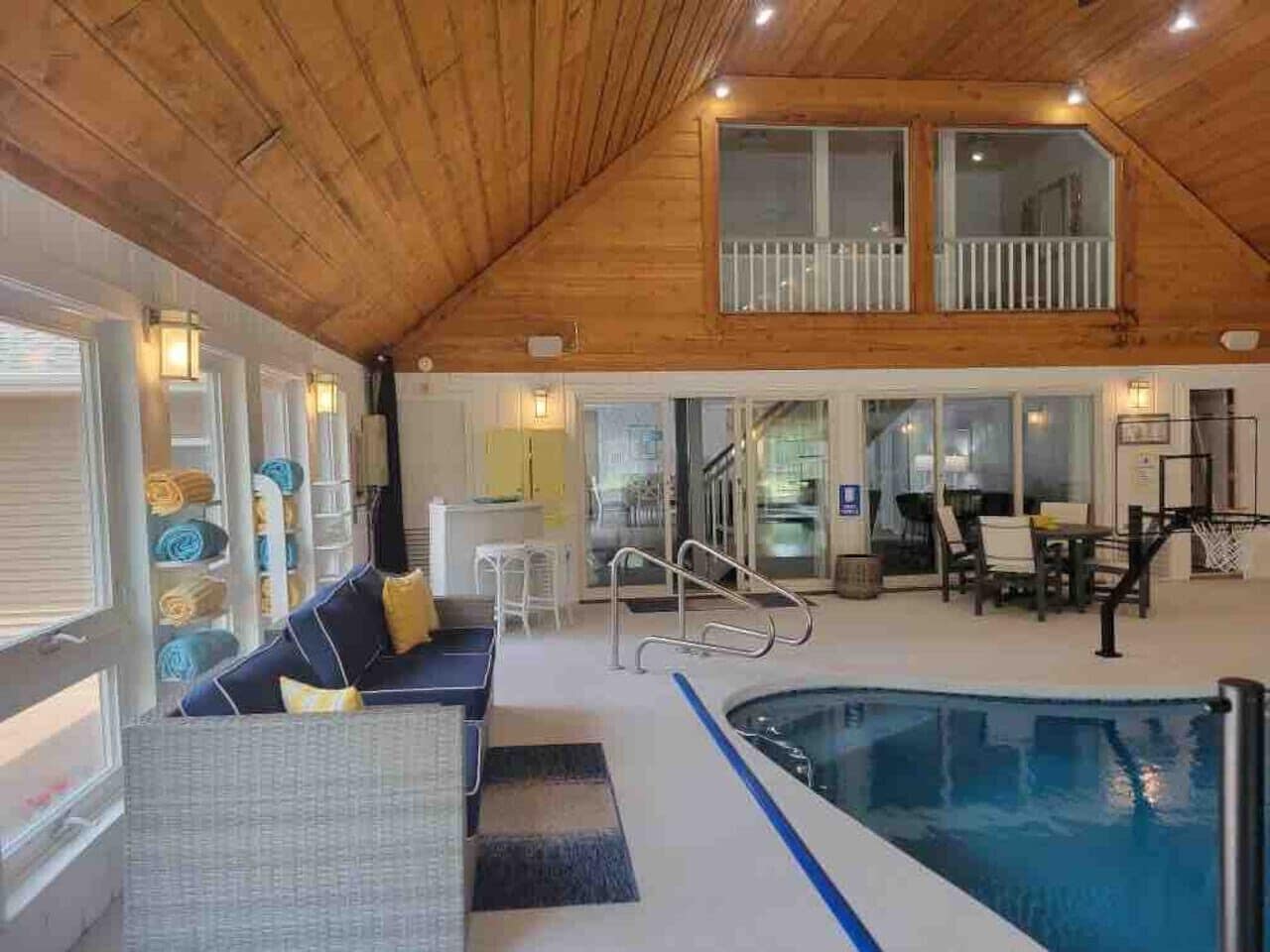 A spacious indoor pool area with a wooden ceiling, a seating area with a sofa and chairs, and a dining table with chairs.