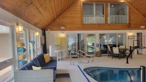 Amplia zona de piscina interior con techo de madera, un área de descanso con sofá y sillones, y una mesa de comedor con sillas.
