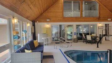 A spacious indoor pool area with a wooden ceiling, a seating area with a sofa and chairs, and a dining table with chairs.