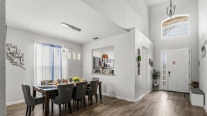 Un coin repas avec une table et des chaises en bois, une cuisine à l’arrière-plan et un couloir menant à une porte.