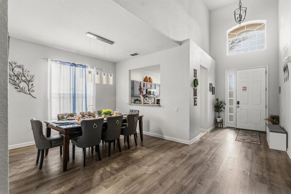 Un coin repas avec une table et des chaises en bois, une cuisine à l’arrière-plan et un couloir menant à une porte.